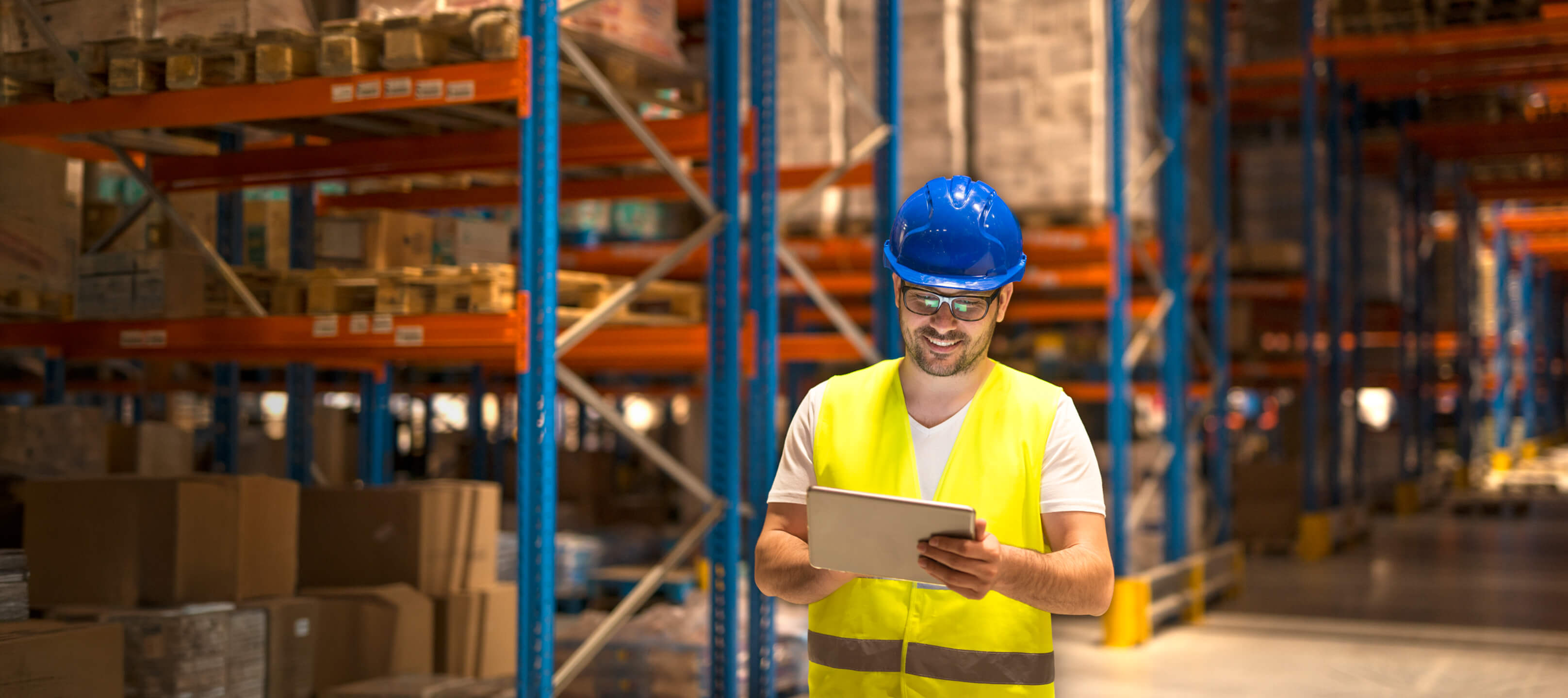 A logistics warehouse worker wearing a blue hard hat and safety vest interacts with an Android tablet, demonstrating enterprise frontline mobile device management (MDM) and rugged fleet tracking software.